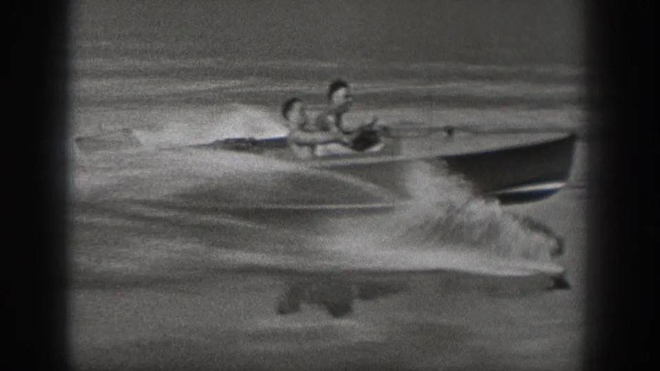 Washington 1940 home-movie frame showing a low motorboat carrying two people, with wake spray spreading across the lake.