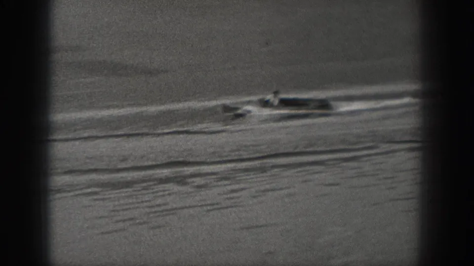 A small boat with two occupants moves across a Washington lake in soft-focus black-and-white 8mm footage.