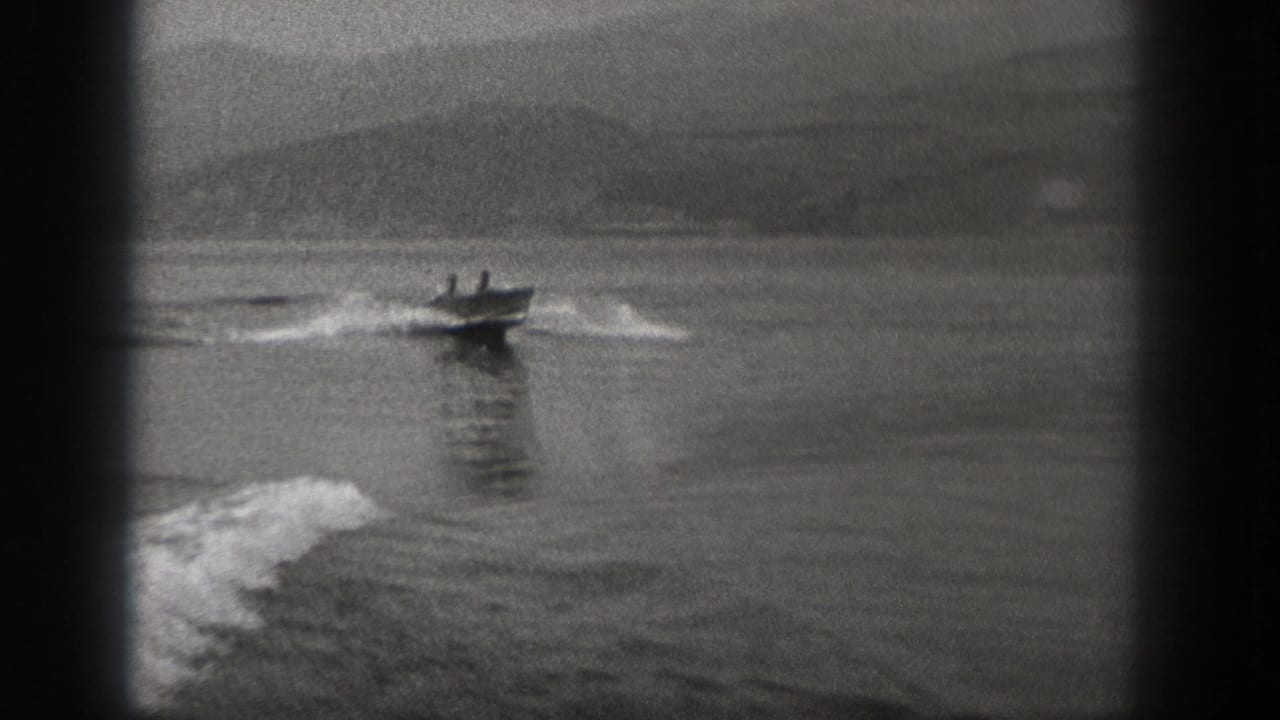 1940 Washington 8mm frame showing two men in a small motorboat speeding across open water with a foamy wake and hazy hills behind.