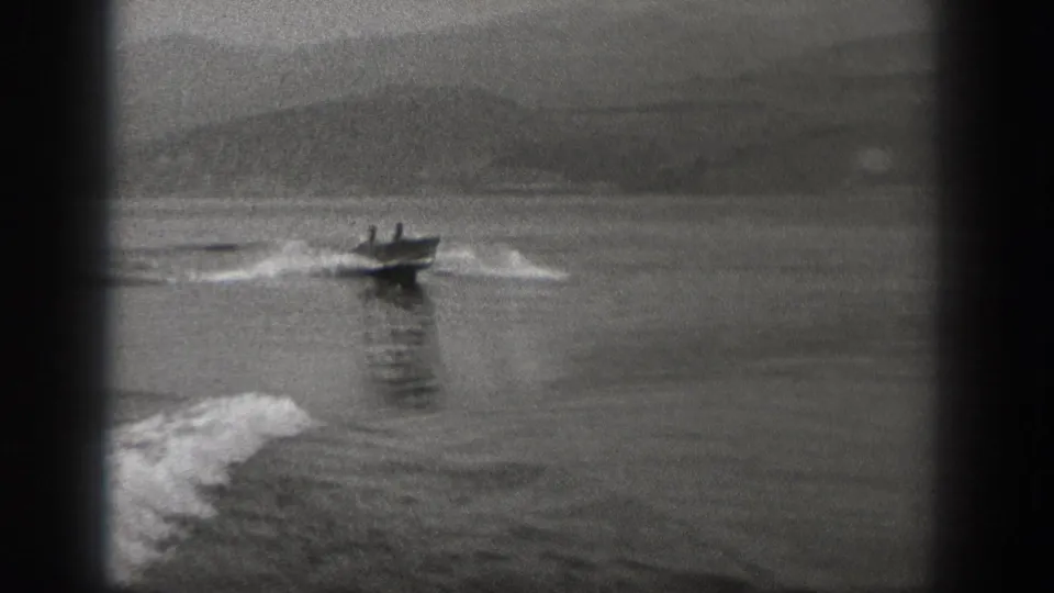 1940 Washington 8mm frame showing two men in a small motorboat speeding across open water with a foamy wake and hazy hills behind.