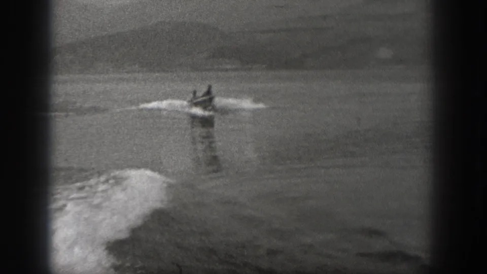 Black-and-white 8mm frame from Washington showing a small motorboat with two men moving fast across a river or lake.