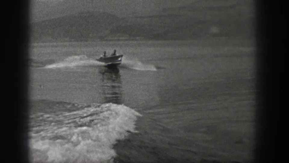 1940 Washington home-movie frame showing two men in a small motorboat on open water, with a larger wake visible in the foreground.