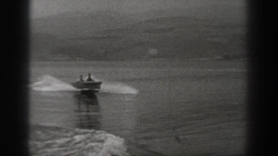 Black-and-white 8mm frame of a small motorboat with two men on Washington water, seen against distant hills and a gray sky.