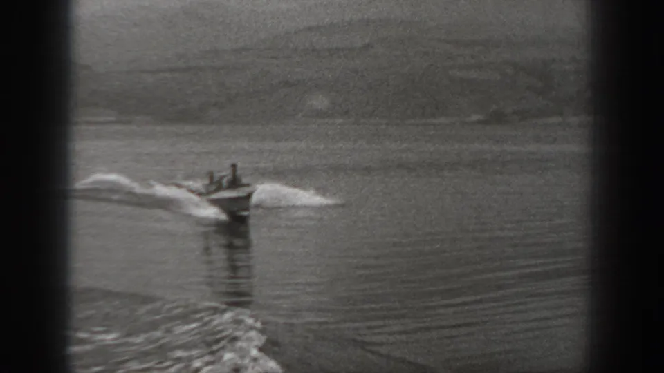 1940 Washington home-movie frame showing two people in a small motorboat crossing calm water with a white wake behind them.