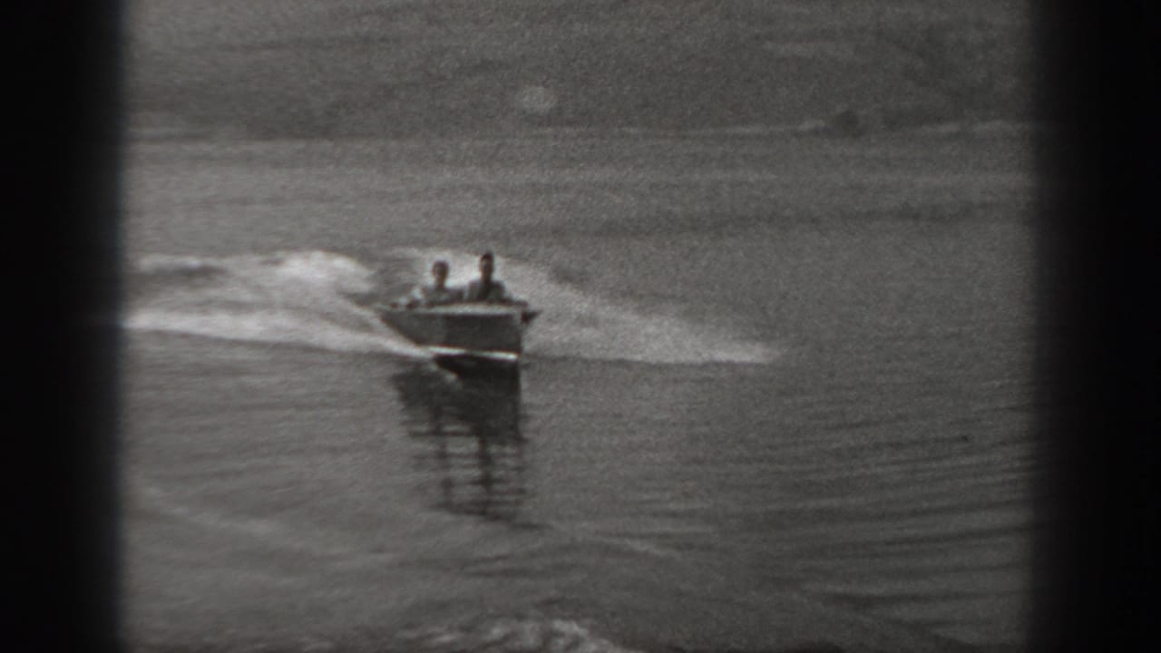 The boat approaches at an angle on a calm Washington lake, with two seated figures visible above the bow and wake spreading behind.