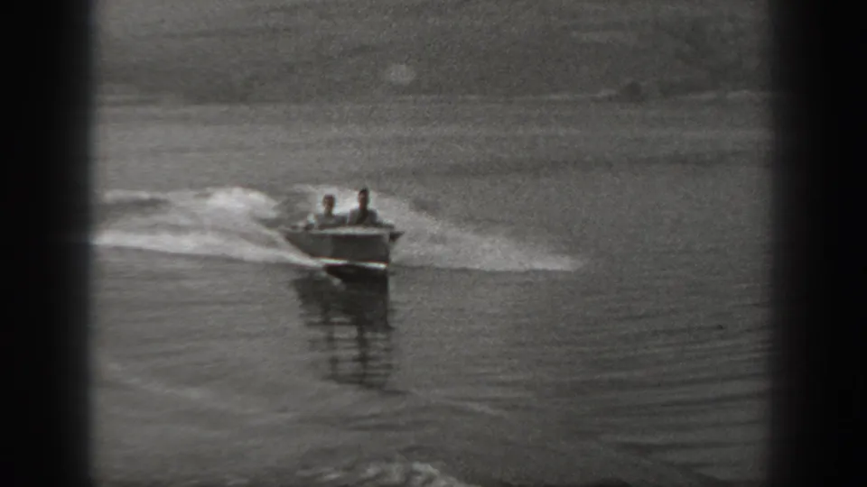 The boat approaches at an angle on a calm Washington lake, with two seated figures visible above the bow and wake spreading behind.