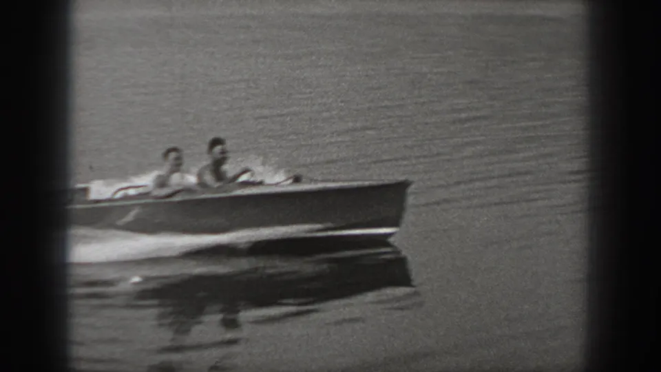 Closer 8mm frame of two people in a small motorboat on calm Washington lake water, with the hull reflected below and wake trailing left.