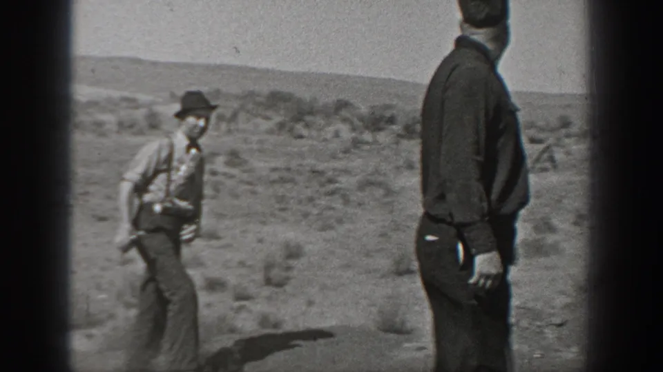 Black-and-white frame of adult men in period clothing walking and standing in a remote Washington desert roadside setting.