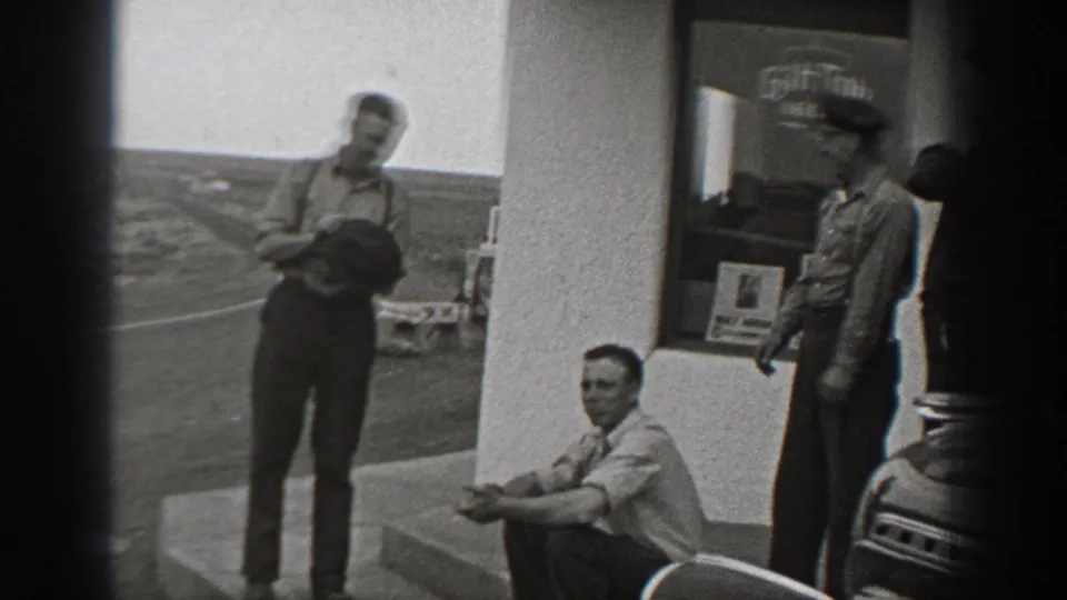 Black-and-white 8mm frame showing adult men outside a roadside cafe or service stop in Washington, with a car nearby.