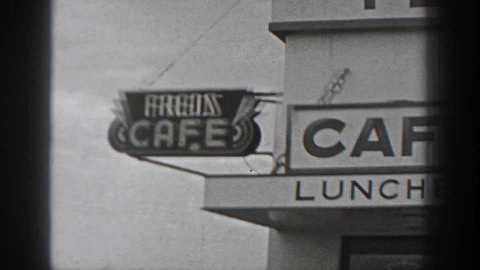 Black-and-white frame showing roadside signage for Fred's Cafe and a larger cafe lunch sign on a building exterior.