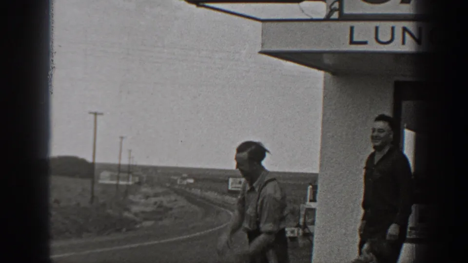 1940 Washington home-movie frame with two men outside Fred's Cafe, roadside terrain, and utility poles in the background.