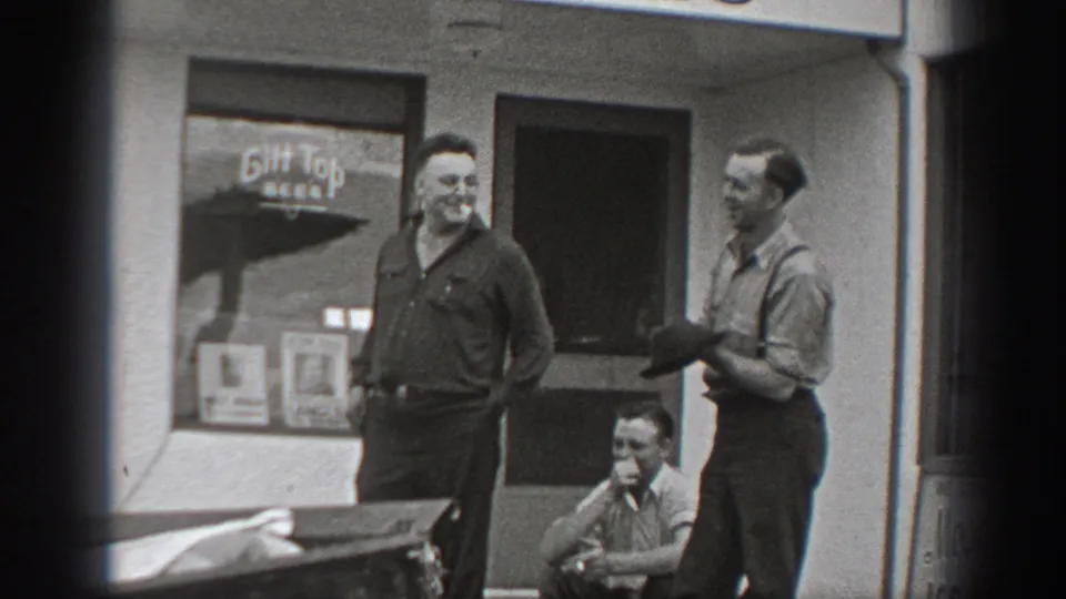 1940 Washington home-movie frame showing men talking outside a storefront, one seated low by the doorway and a Gilt Top Beer sign visible behind them.