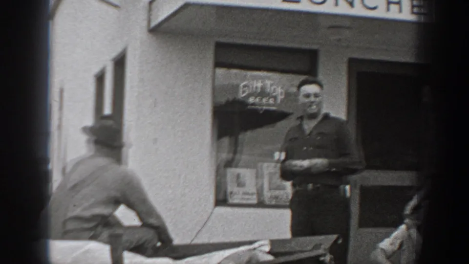 1940 Washington home-movie frame with a man standing near the beer sign window as another adult passes across the foreground.