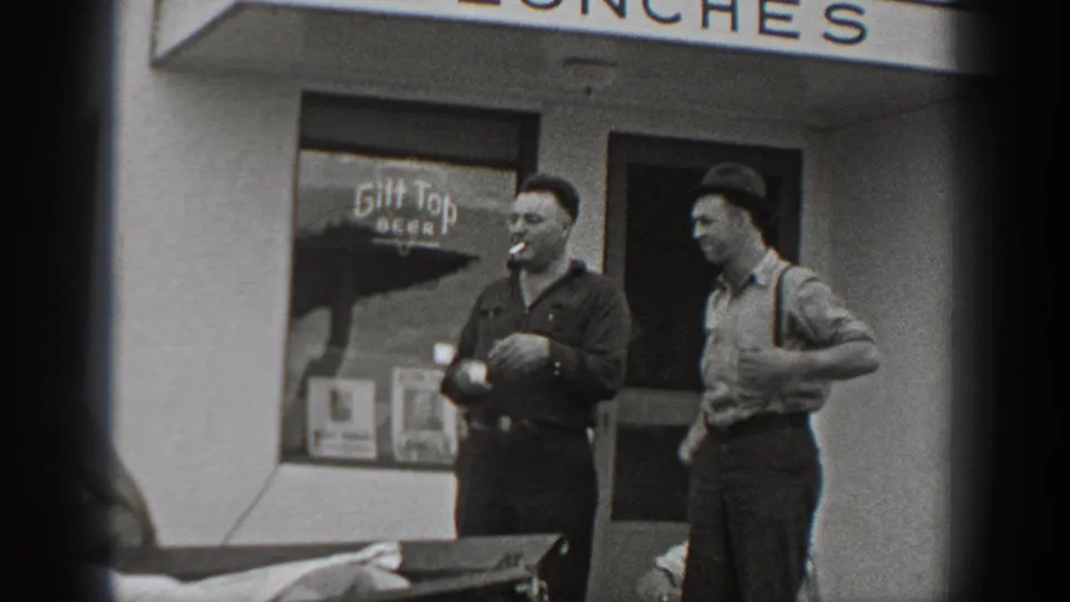 1940 Washington frame showing two adult men beside a storefront doorway, with one smoking and beer advertising visible in the window.