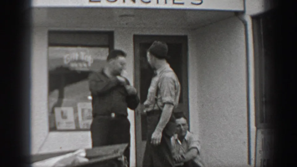 Black-and-white 1940 Washington storefront scene with two adult men conversing and a seated man near a window with beer signage.