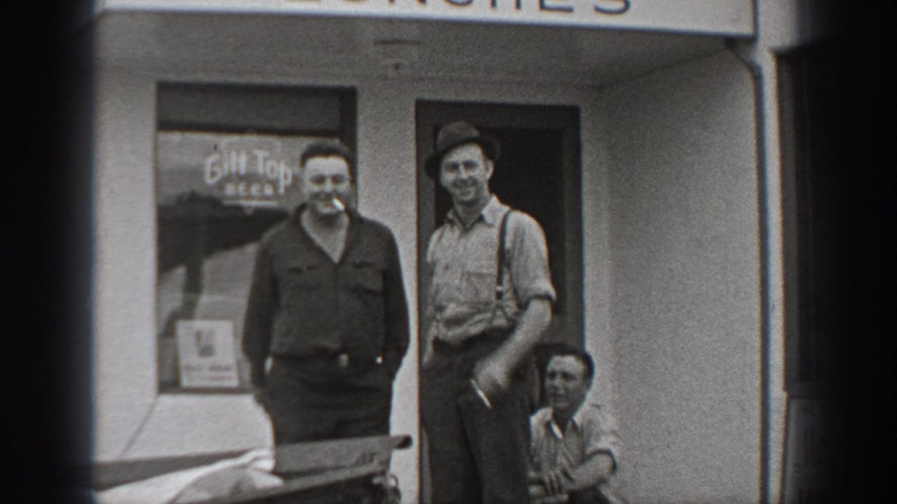 Two standing men and a seated or crouching man outside a storefront with beer signage.