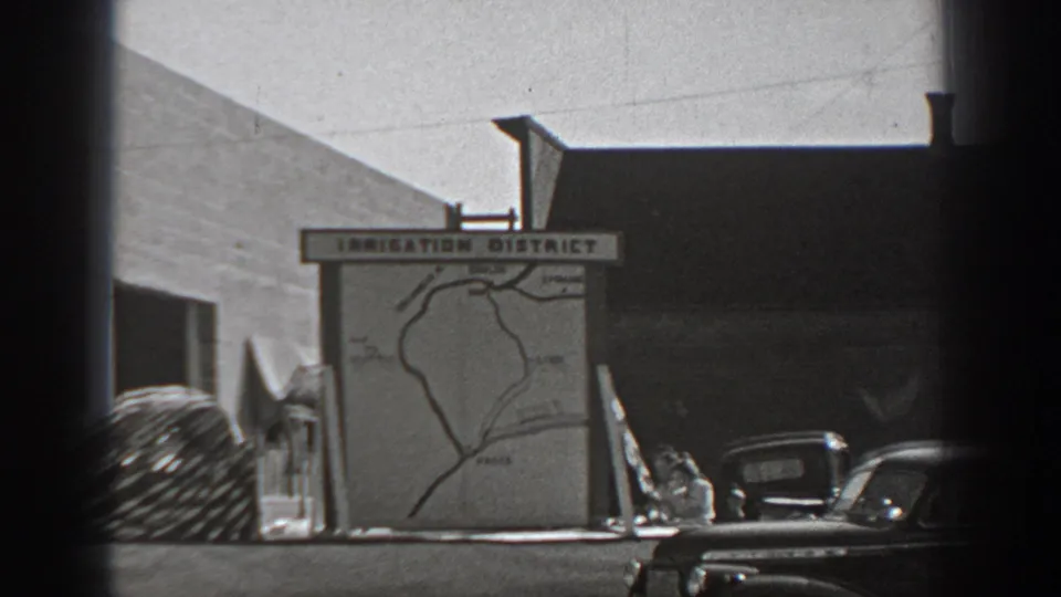 1940 Washington home-movie frame showing an Irrigation District map sign near parked cars and a building wall.