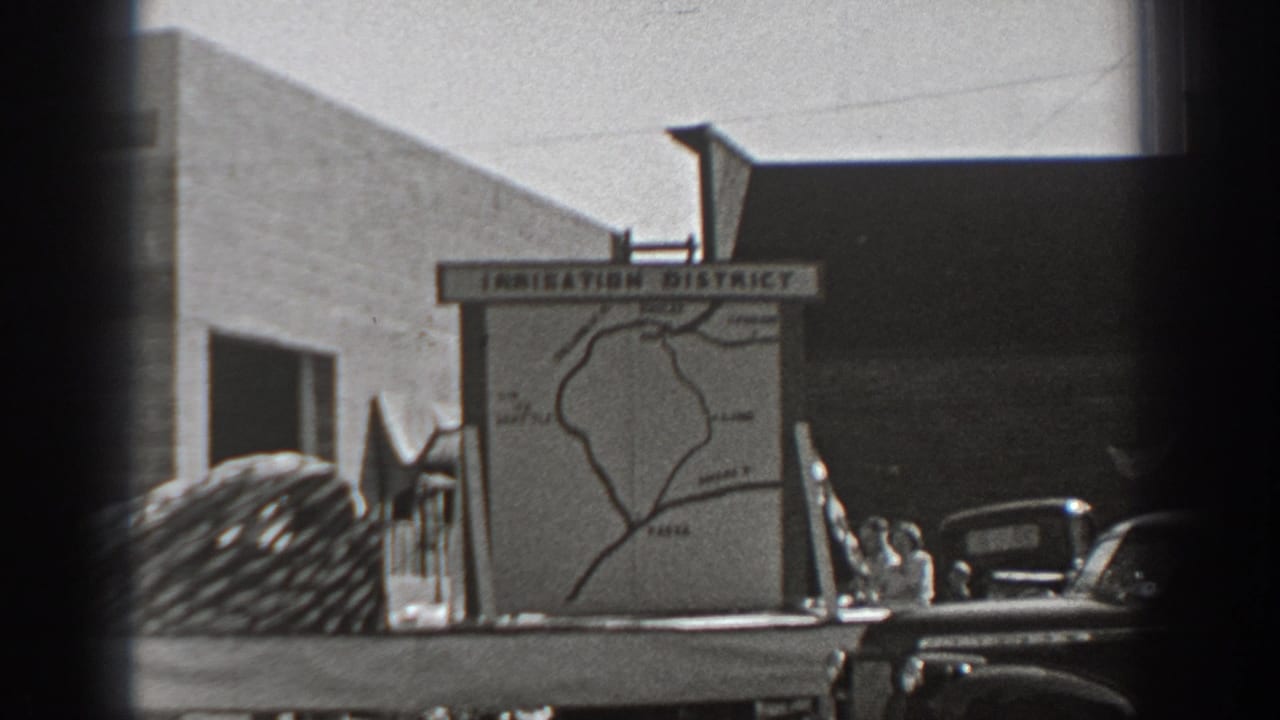 1940 Washington home-movie frame showing an irrigation district map board with parked trucks, a coiled load, and low buildings in bright daylight.