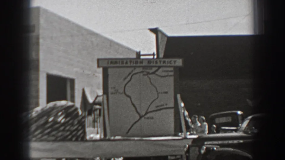 1940 Washington home-movie frame showing an irrigation district map board with parked trucks, a coiled load, and low buildings in bright daylight.