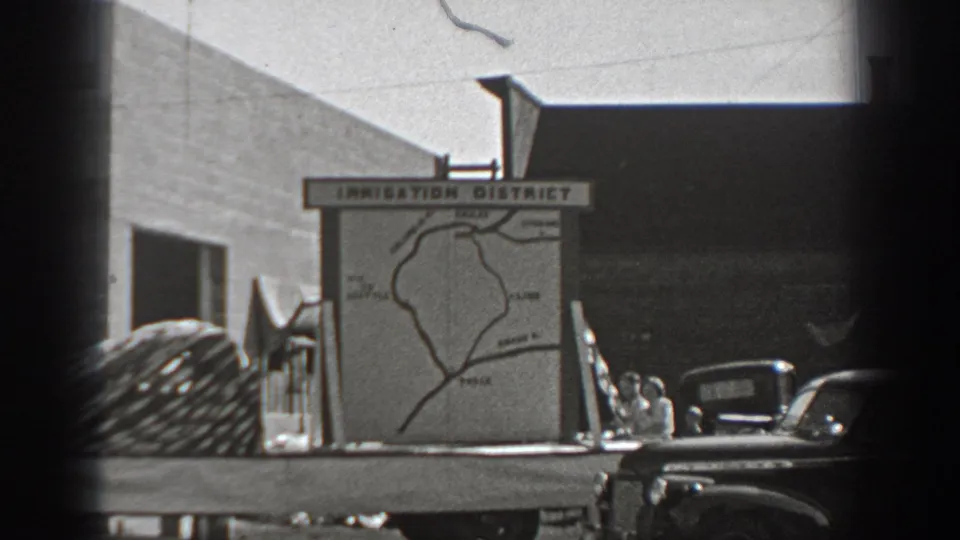 The irrigation district board remains centered, with vintage vehicles, small figures, and a large building visible behind it.