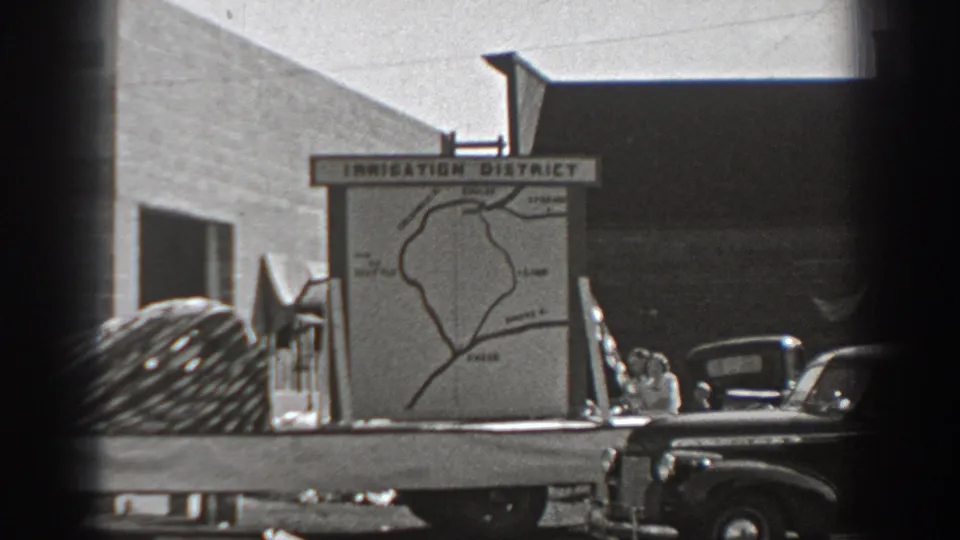 A black-and-white 8mm frame shows the irrigation district map board, a large coil on the truck bed, and older vehicles beside simple buildings.