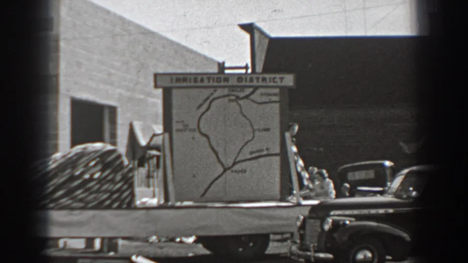 1940 Washington roadside view of an Irrigation District map sign on a flatbed truck, with period cars and a plain building wall nearby.