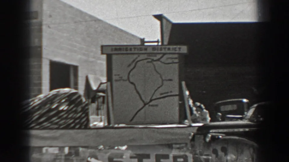 The Irrigation District map sign remains centered on the truck bed, with cars, a building facade, and dark 8mm frame edges visible.