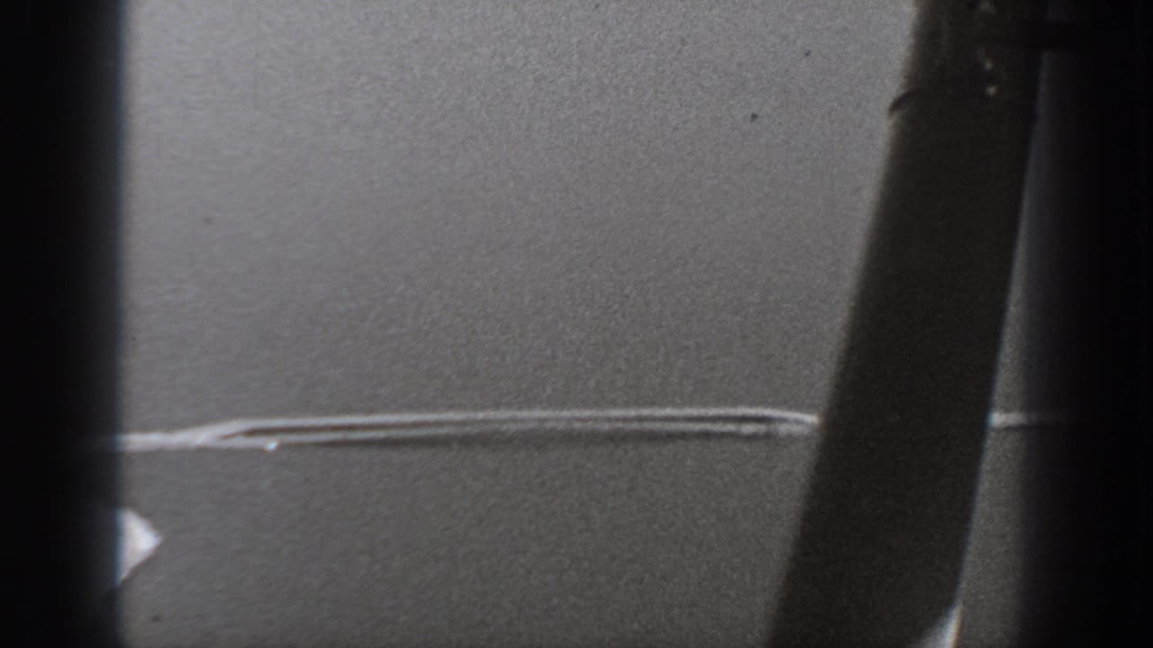 1940 Washington home-movie frame looking through a dark aircraft window toward a soft gray surface crossed by a thin bright streak.