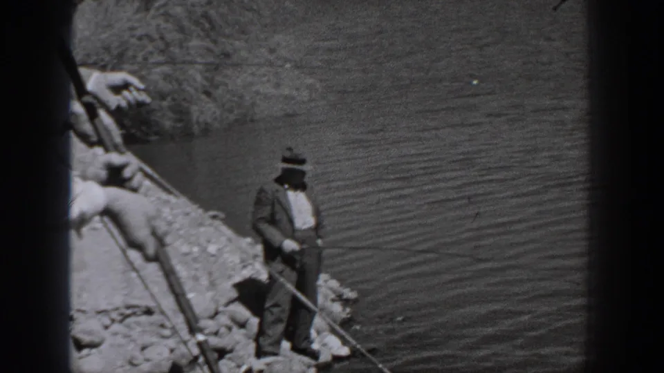 Man in a hat and suit-like clothing fishing from a rocky riverbank in Washington, shown in 1940 black-and-white 8mm footage.