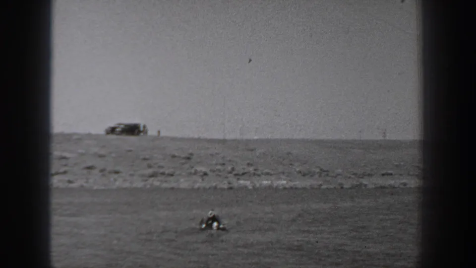 Wide Washington 1940 home-movie view of low shoreline, a distant vehicle, and a small figure or object in the water.