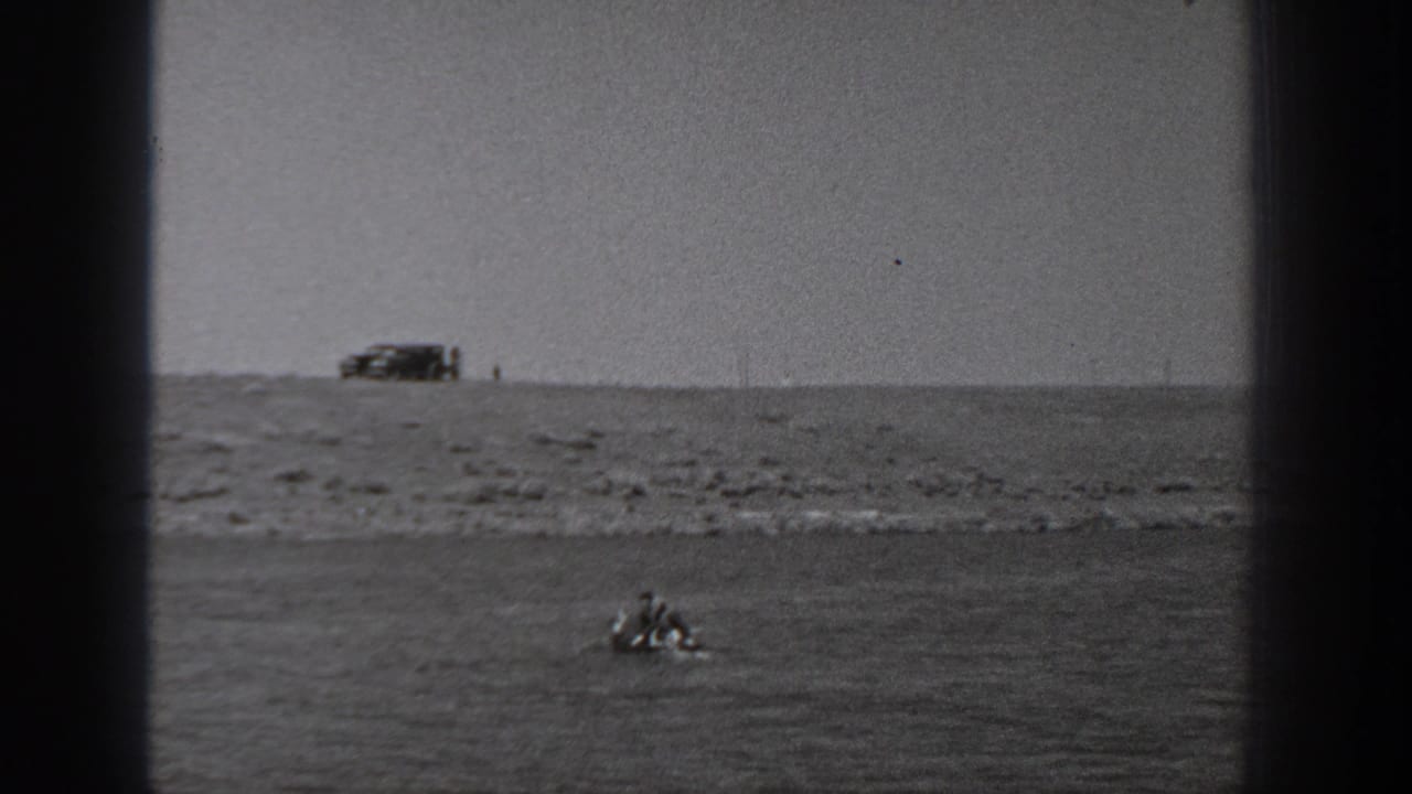 1940 Washington 8mm frame showing two people in a small boat near a rocky shoreline with a vehicle parked on the bluff above.