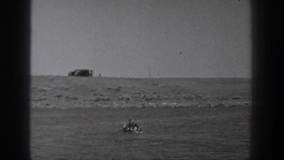 1940 Washington 8mm frame showing two people in a small boat near a rocky shoreline with a vehicle parked on the bluff above.