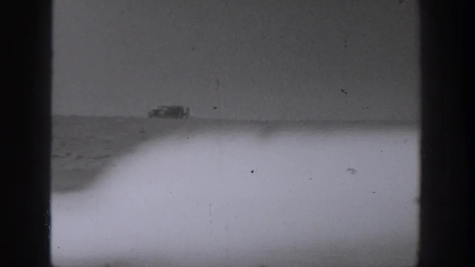 Partially blocked 1940 Washington home-movie frame showing water and a distant shoreline vehicle, with the boat largely hidden by exposure or obstruction.