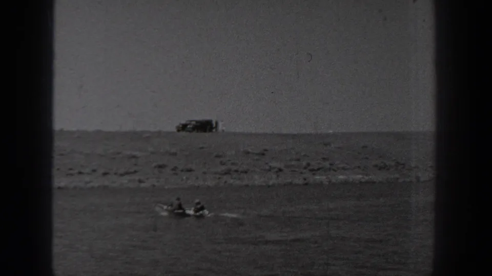 1940 Washington 8mm frame showing the small boat farther across the water beneath a rocky shoreline and distant parked vehicle.