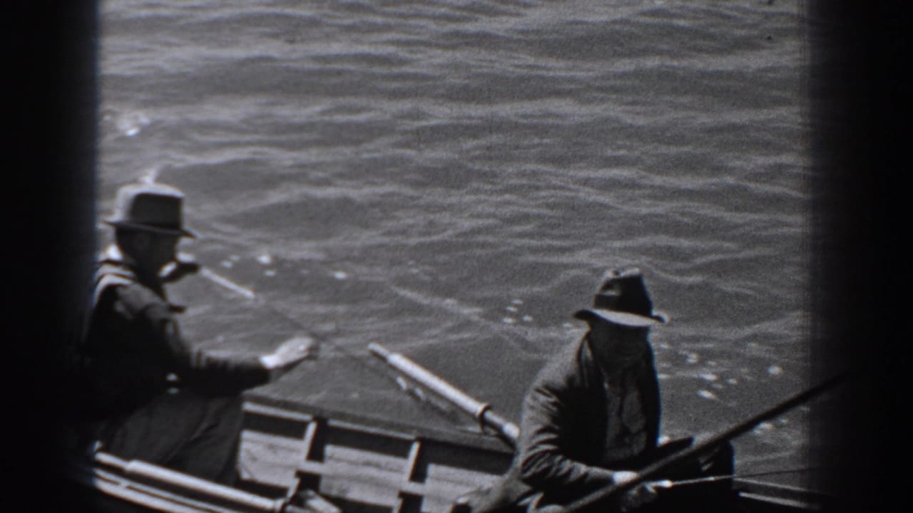 1940 Washington home-movie frame showing two men in dark coats and brimmed hats fishing from a small rowboat on choppy water.
