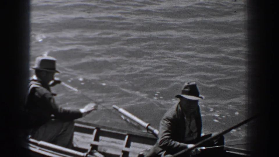 1940 Washington home-movie frame showing two men in dark coats and brimmed hats fishing from a small rowboat on choppy water.