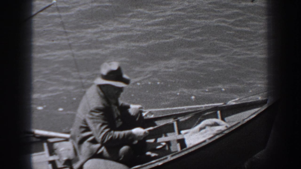 A man seated in a rowboat holds a fishing rod as water fills the background.