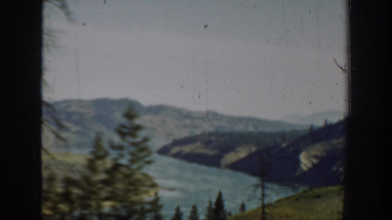 1940s Washington 8mm travel view of a mountain waterway, green slopes, and foreground evergreens framed by a vehicle or train window.
