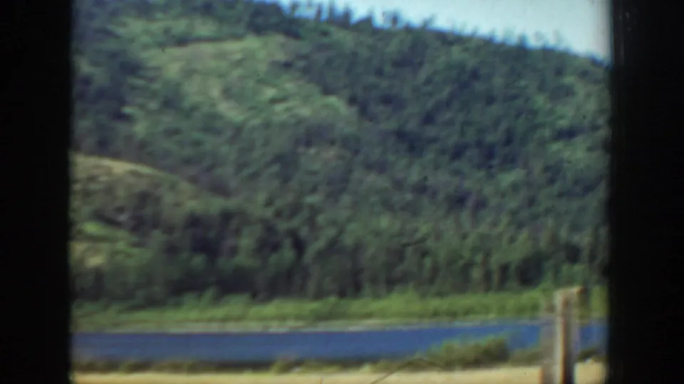 1940s Washington scenic frame showing blue water, a forested hillside, green shoreline, and fence posts near the foreground.