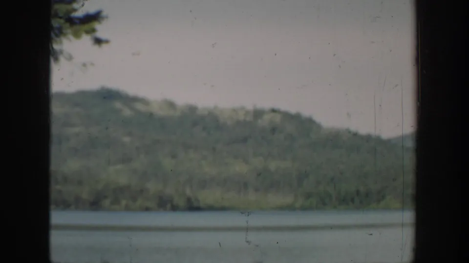 1940 Washington home-movie frame of a lake bordered by wooded hills, with light scratches and soft color fading.