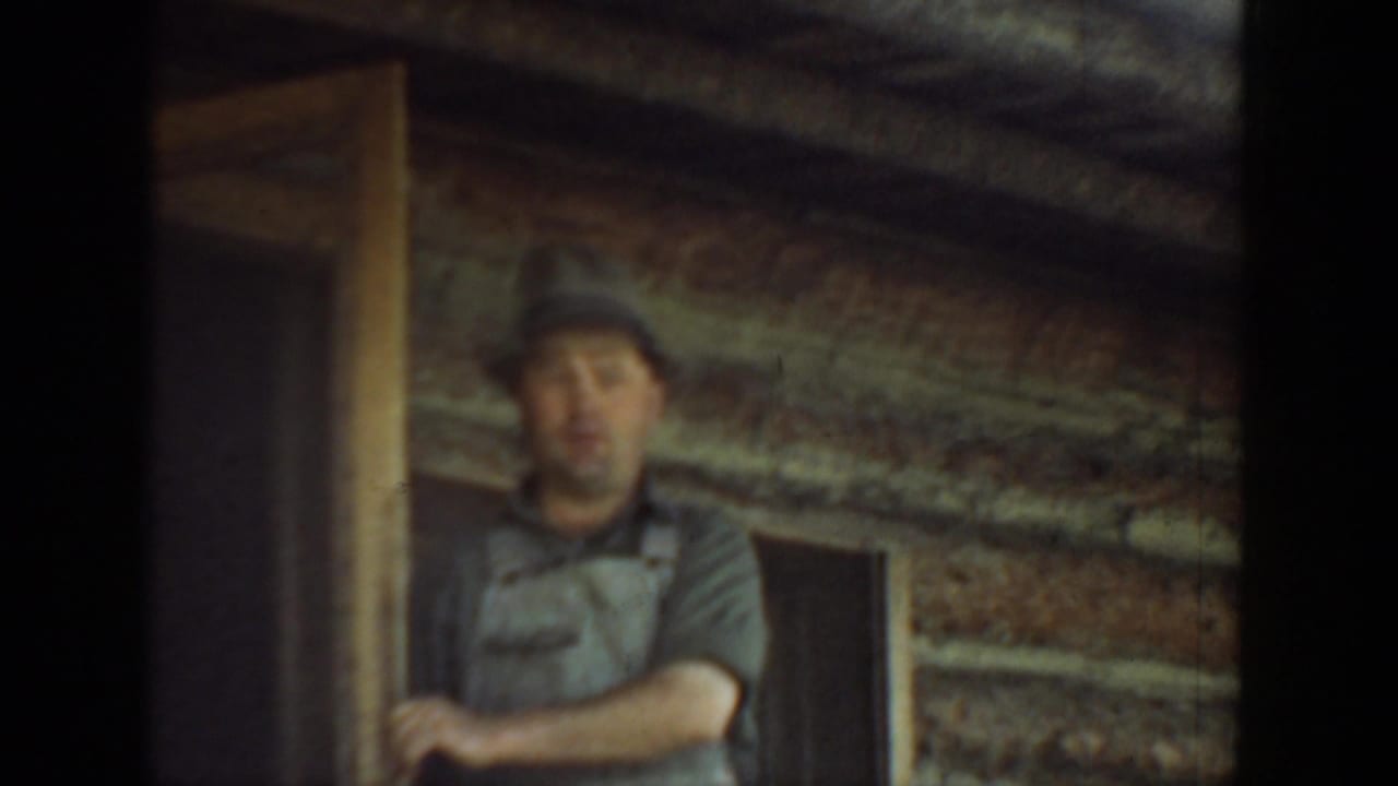 Adult man in a cap and work shirt stands in a log cabin doorway in Washington 1940 home-movie footage.