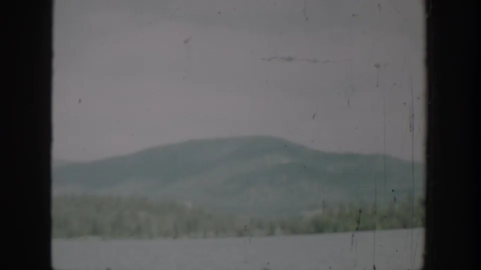 1940 Washington 8mm frame with pale lake water, a low forested ridge, and muted hills in soft focus.