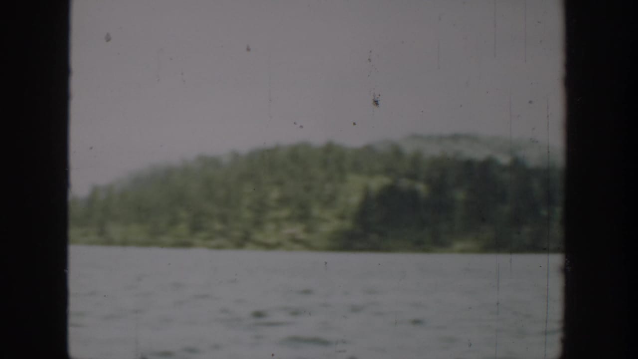 1940 Washington 8mm frame with open lake water, tree-covered shoreline, low hills, pale sky, and visible film wear.