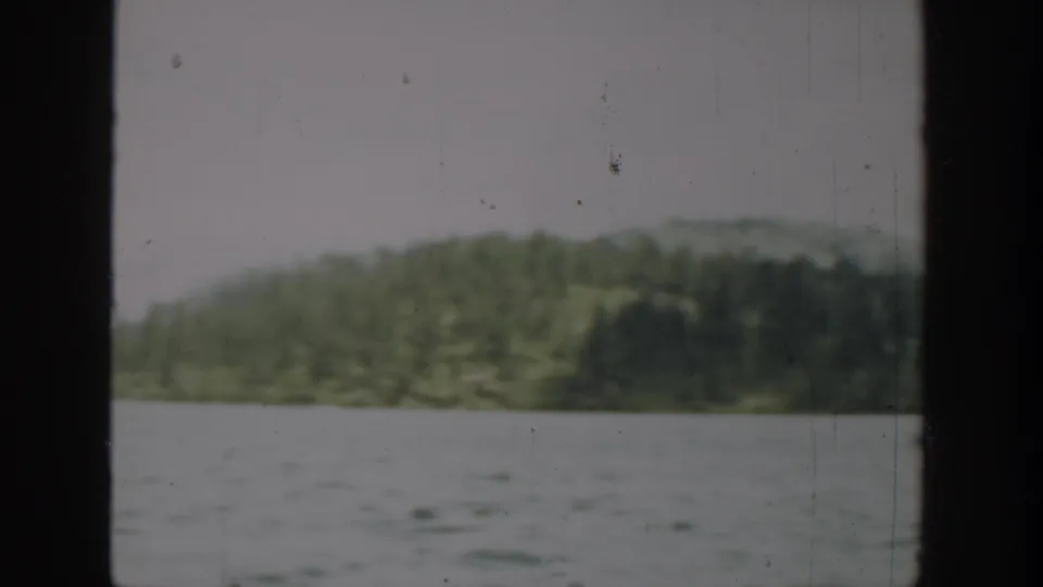 1940 Washington 8mm frame with open lake water, tree-covered shoreline, low hills, pale sky, and visible film wear.