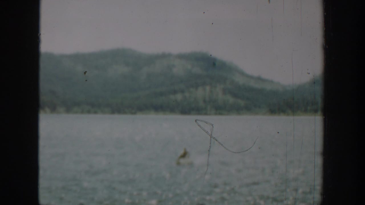 1940 Washington home-movie frame of a distant small boat on a lake, viewed through a dark window frame with wooded hills in the background.