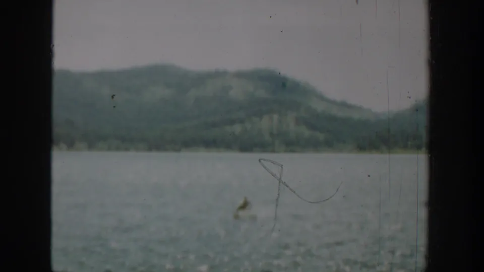1940 Washington home-movie frame of a distant small boat on a lake, viewed through a dark window frame with wooded hills in the background.