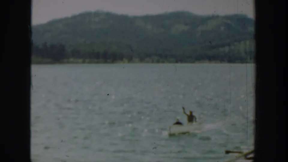 Two people ride in a small boat on a Washington lake, with one person raising an arm toward the camera in the 1940 home-movie frame.