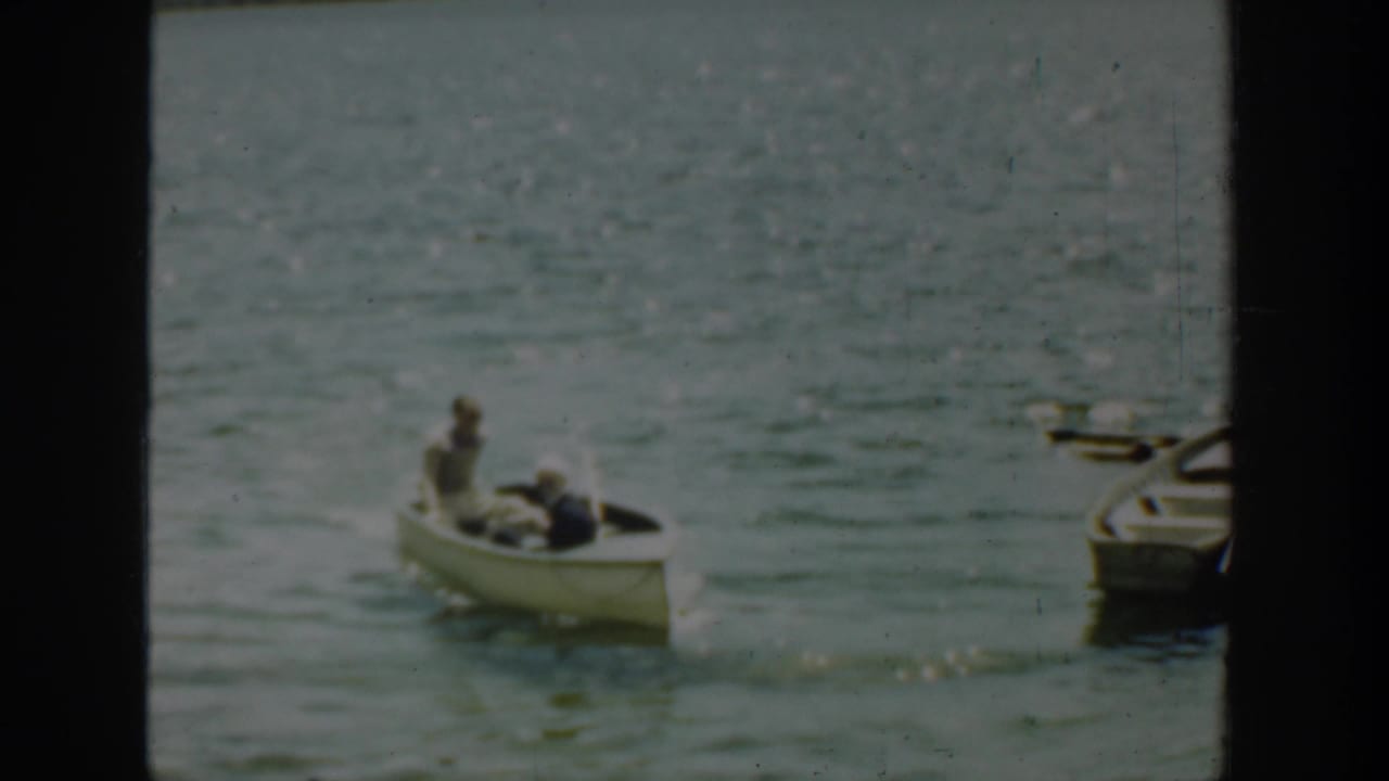 A rowboat with two people moves across a lake near other boats at the right edge.