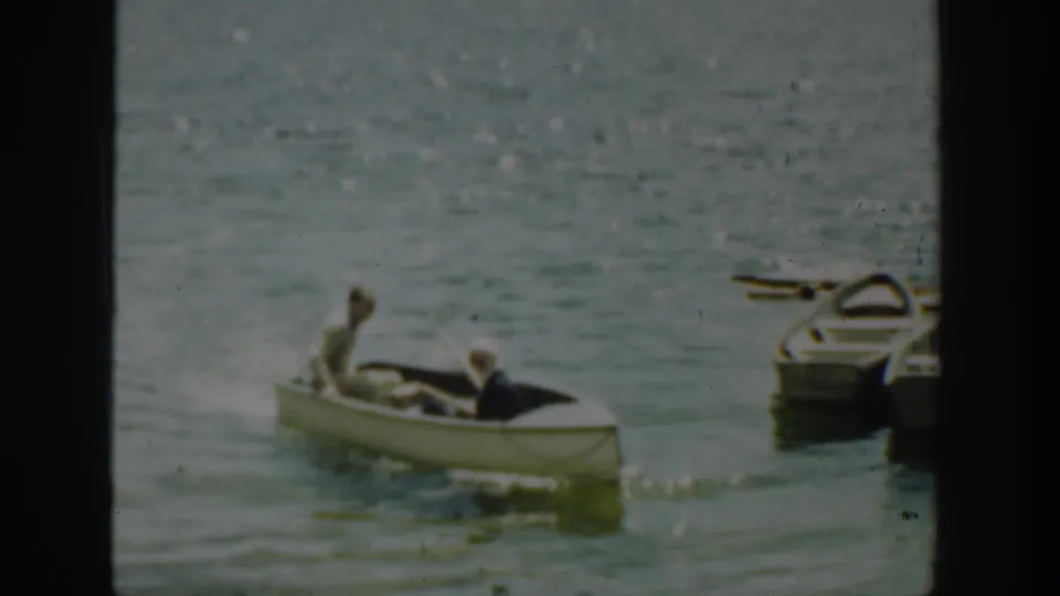 A man rows a small white boat with another passenger seated ahead, passing docked rowboats in Washington lake footage.
