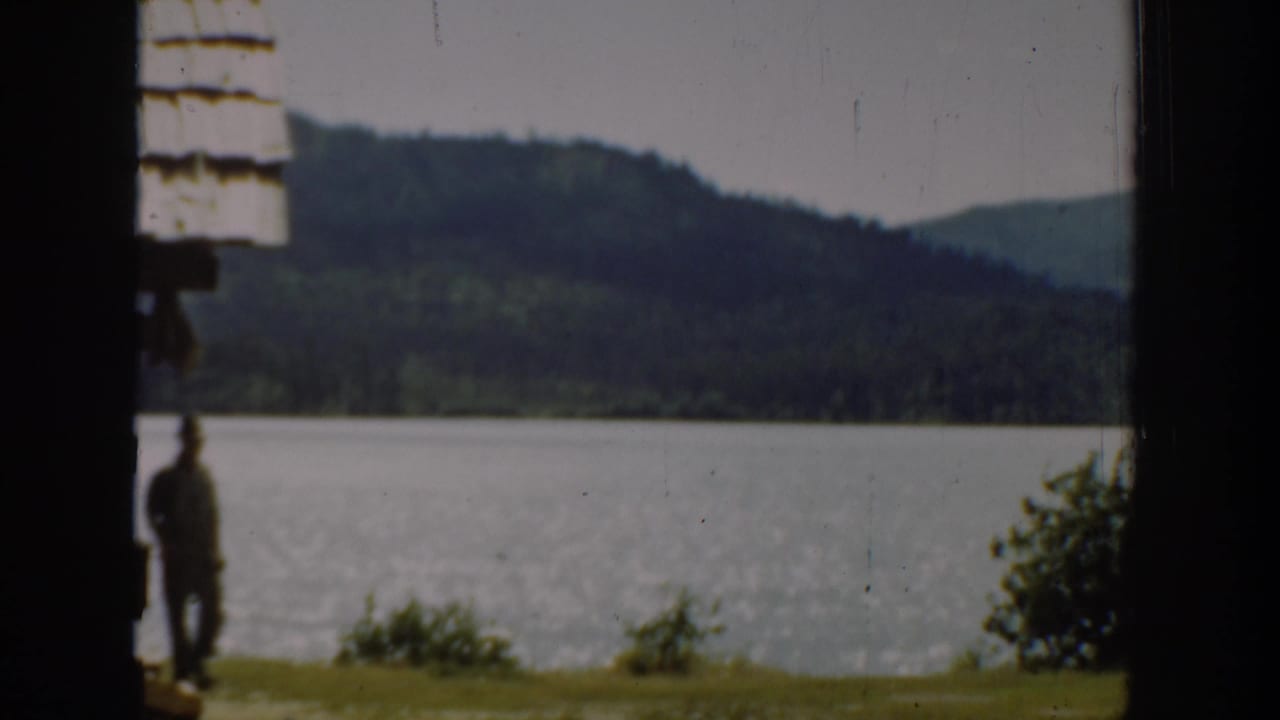 Man standing near a lakeshore, seen through a dark vertical frame with hills behind him.